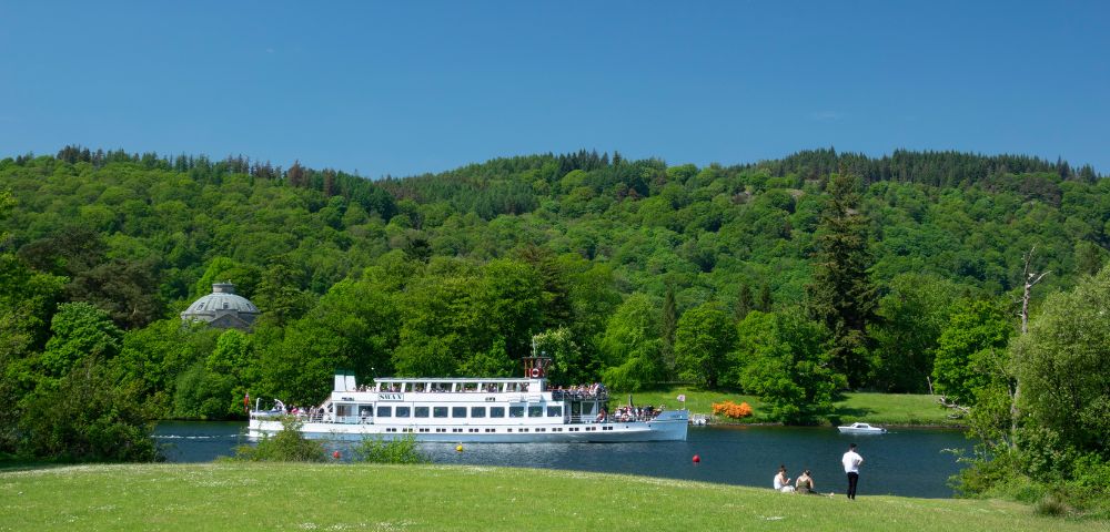 A serene landscape featuring a white boat cruising on a tranquil river, surrounded by lush green hills and trees. People sit on the grassy bank under a clear blue sky, evoking a peaceful, leisurely vibe.