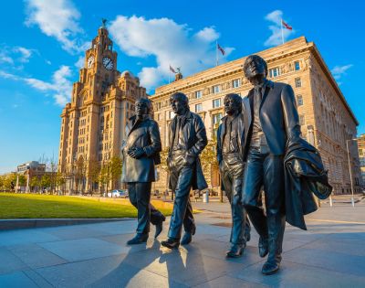 Bronze statues of four men walk together, set against the backdrop of a historic building and clear blue sky in Liverpool. The mood is vibrant and iconic.