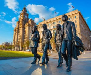 Bronze statues of four men walk together, set against the backdrop of a historic building and clear blue sky in Liverpool. The mood is vibrant and iconic.