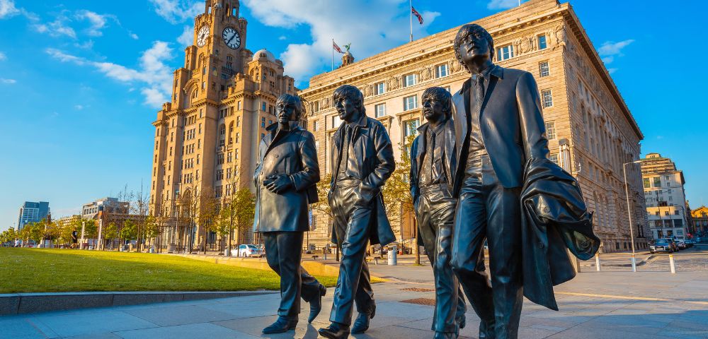 Bronze statues of four men walk together, set against the backdrop of a historic building and clear blue sky in Liverpool. The mood is vibrant and iconic.