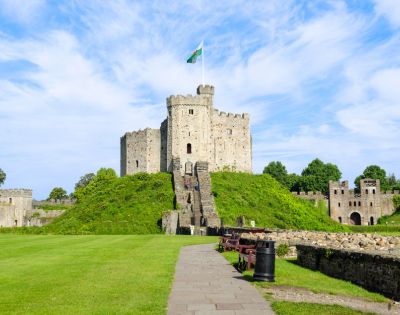 A historic stone castle atop a grassy hill with a Welsh flag flying high. Surrounded by lush greenery, benches, and a partly cloudy sky.