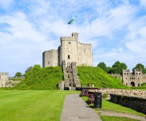 A historic stone castle atop a grassy hill with a Welsh flag flying high. Surrounded by lush greenery, benches, and a partly cloudy sky.