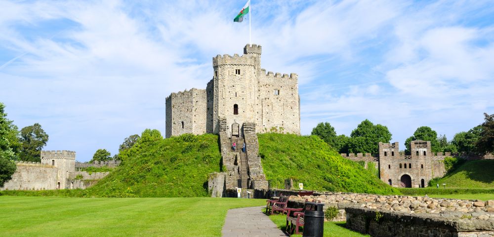 A historic stone castle atop a grassy hill with a Welsh flag flying high. Surrounded by lush greenery, benches, and a partly cloudy sky.