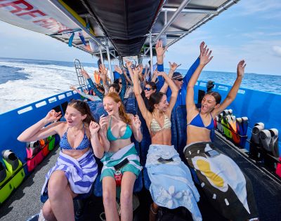 Group of joyful people in swimsuits on a boat, wrapped in towels, raising their arms. Bright day, ocean in background; conveys excitement and fun.