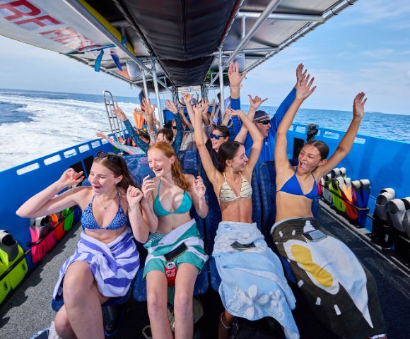 Group of joyful people in swimsuits on a boat, wrapped in towels, raising their arms. Bright day, ocean in background; conveys excitement and fun.