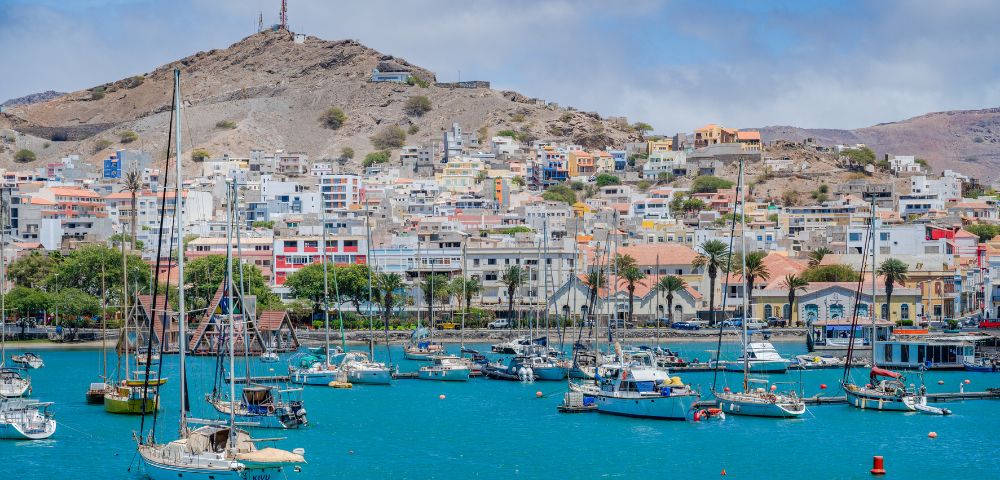 A vibrant harbor scene with sailboats floating on turquoise water and colorful houses on a hillside in the background under a partly cloudy sky.