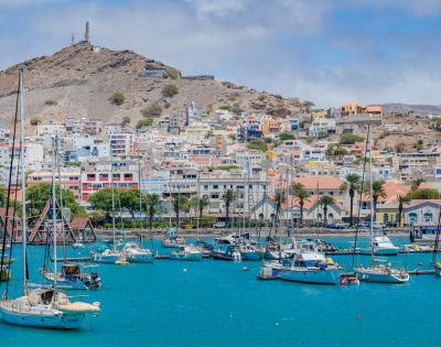 A vibrant harbor scene with sailboats floating on turquoise water and colorful houses on a hillside in the background under a partly cloudy sky.
