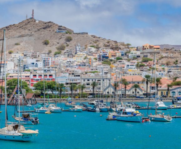 A vibrant harbor scene with sailboats floating on turquoise water and colorful houses on a hillside in the background under a partly cloudy sky.