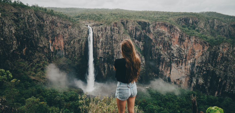 A woman stands in front of a cascading waterfall, in Cairns surrounded by lush greenery and mist rising from the water.