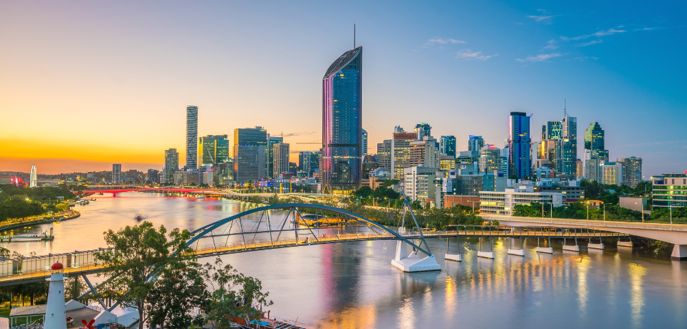 Cityscape of Brisbane, Australia at sunset, showing modern skyscrapers, bridges, and the Brisbane River in the foreground.