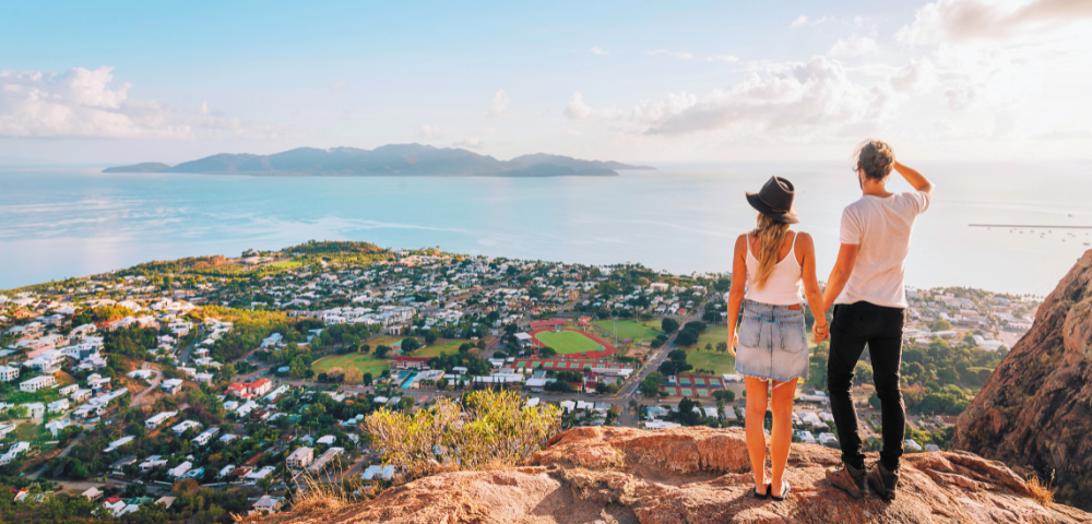 A couple holds hands while gazing at a scenic coastal view from a mountaintop, overlooking a lush town and serene ocean.