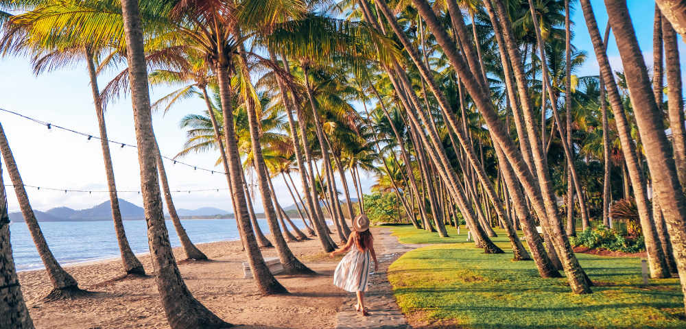 A woman in a sun hat walks along a palm-lined path by the beach, with soft waves and gentle hills in the background.