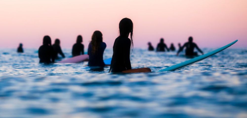 Silhouetted surfers awaiting waves in calm waters at dusk, with a pastel sky reflecting the tranquil evening atmosphere.