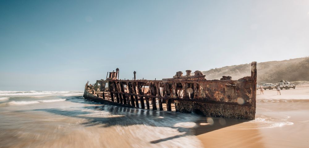 Rusty shipwreck partially submerged on a sandy beach, with gentle waves lapping and clear skies in the background.