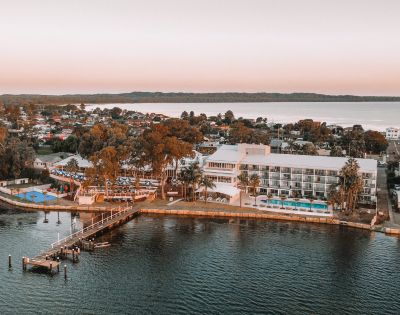 Aerial view of a waterfront resort at sunset, featuring modern buildings, palm trees, a dock, and calm waters. The scene is serene and picturesque.