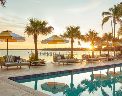 Poolside scene at sunset, with yellow-striped umbrellas, loungers, and palm trees reflecting in the water. A peaceful lake is seen in the background.