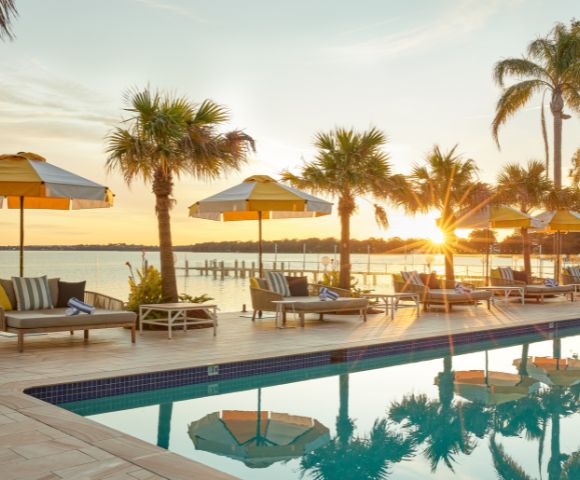 Poolside scene at sunset, with yellow-striped umbrellas, loungers, and palm trees reflecting in the water. A peaceful lake is seen in the background.