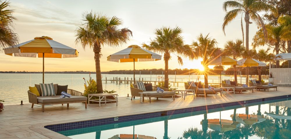 Poolside scene at sunset, with yellow-striped umbrellas, loungers, and palm trees reflecting in the water. A peaceful lake is seen in the background.