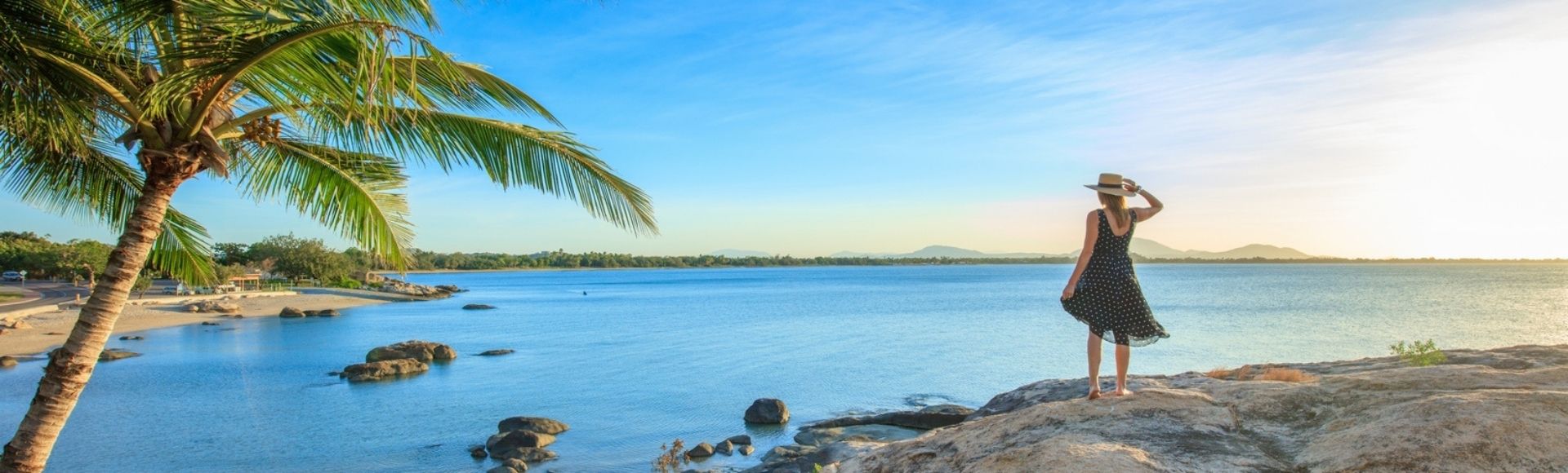 A woman stands on a rock, gazing at the ocean and palm trees in the distance.