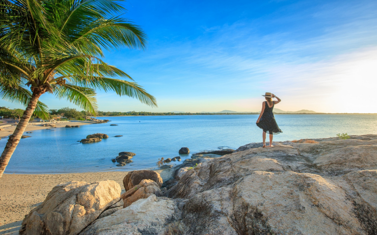 A woman stands on a rock, gazing at the ocean and palm trees in the distance.