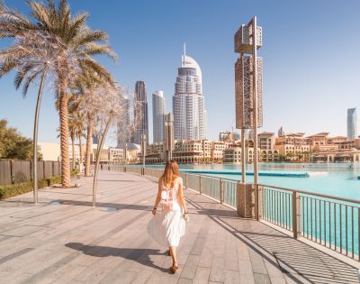 A woman in a white dress walks on a sunny waterfront promenade with skyscrapers and palm trees in the background, conveying a serene urban vibe.