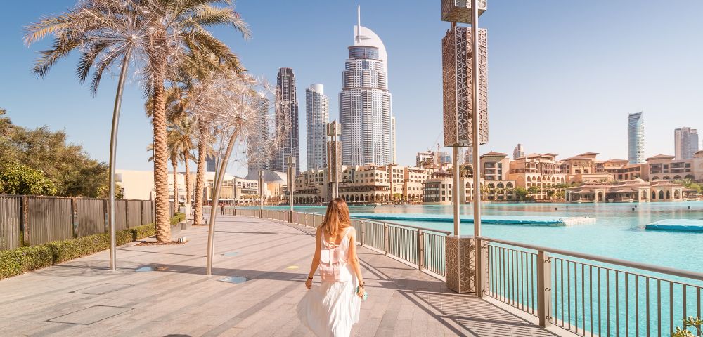 A woman in a white dress walks on a sunny waterfront promenade with skyscrapers and palm trees in the background, conveying a serene urban vibe.
