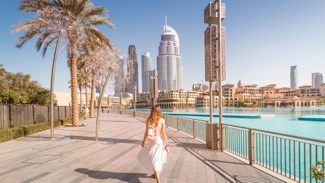 A woman in a white dress walks on a sunny waterfront promenade with skyscrapers and palm trees in the background, conveying a serene urban vibe.
