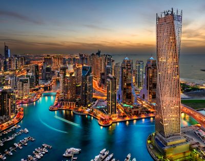 Aerial view of a vibrant cityscape at dusk with illuminated skyscrapers, a winding waterway, and a serene sky transitioning from sunset to night.