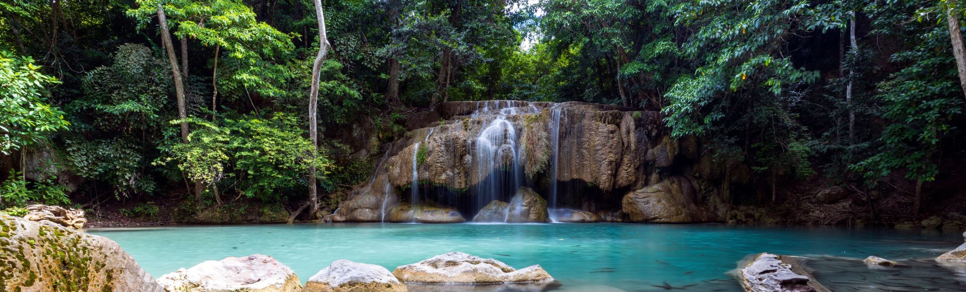 A serene jungle scene featuring a turquoise pond with smooth stones, a gentle waterfall cascading over rocks, and lush greenery surrounding it in Erawan National Park, Thailand.