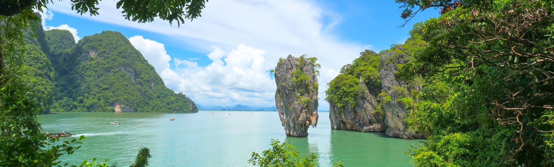 Serene coastal scene in Phang Nga Bay featuring towering limestone islets emerging from tranquil waters against a bright blue sky, framed by lush greenery.