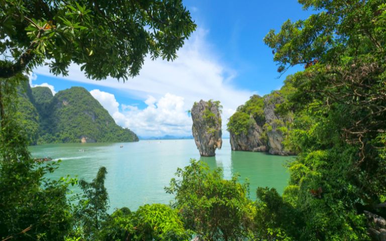 Serene coastal scene in Phang Nga Bay featuring towering limestone islets emerging from tranquil waters against a bright blue sky, framed by lush greenery.