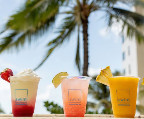 Three colorful cocktails with fruit garnishes sit against a tropical backdrop. A blurred palm tree and building are visible under a clear blue sky.