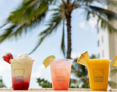 Three colorful cocktails with fruit garnishes sit against a tropical backdrop. A blurred palm tree and building are visible under a clear blue sky.