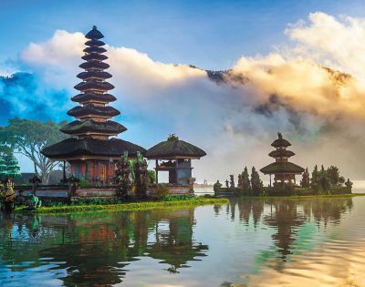 A serene lake scene with a Balinese temple featuring tiered roofs, reflected on the water. Surrounded by lush trees and dramatic clouds under a vibrant sky.