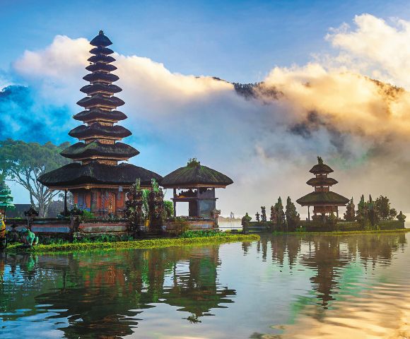 A serene lake scene with a Balinese temple featuring tiered roofs, reflected on the water. Surrounded by lush trees and dramatic clouds under a vibrant sky.