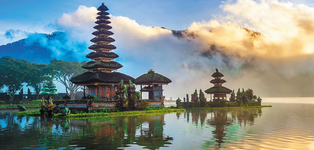 A serene lake scene with a Balinese temple featuring tiered roofs, reflected on the water. Surrounded by lush trees and dramatic clouds under a vibrant sky.