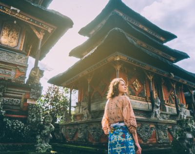 Woman in traditional attire stands before ornate Balinese temple under clear skies. Sunlight softly illuminates the intricate carvings, creating a serene atmosphere.
