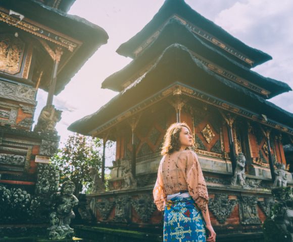 Woman in traditional attire stands before ornate Balinese temple under clear skies. Sunlight softly illuminates the intricate carvings, creating a serene atmosphere.