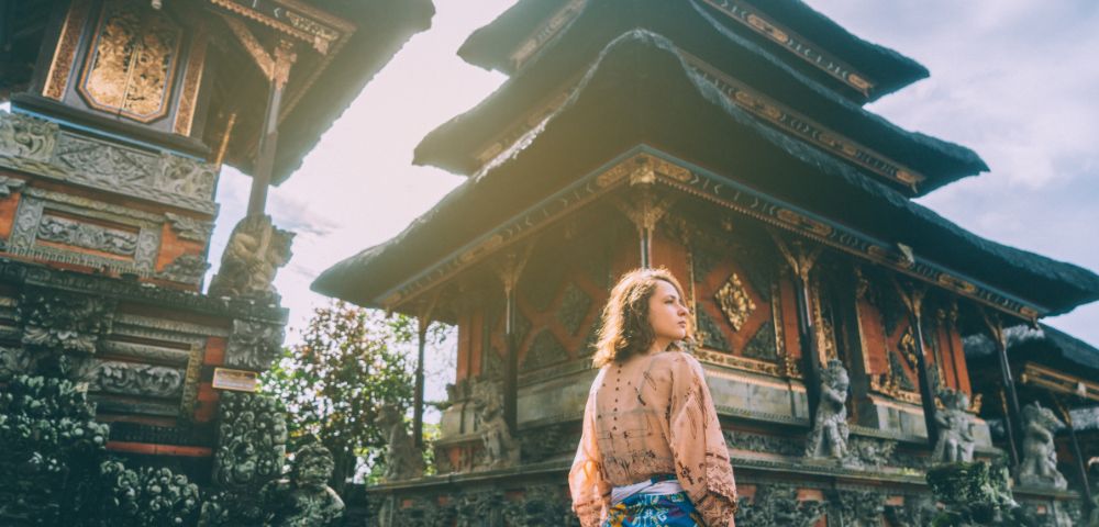 Woman in traditional attire stands before ornate Balinese temple under clear skies. Sunlight softly illuminates the intricate carvings, creating a serene atmosphere.