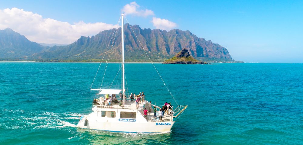 A sailing boat with passengers on bright turquoise waters, set against lush green mountains under a clear blue sky.