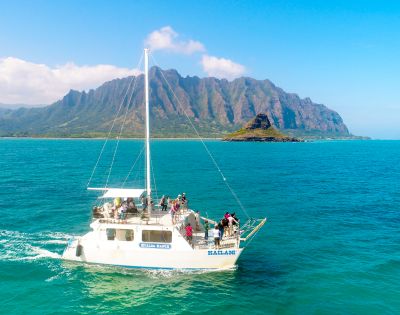 A sailing boat with passengers on bright turquoise waters, set against lush green mountains under a clear blue sky.
