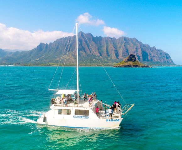 A sailing boat with passengers on bright turquoise waters, set against lush green mountains under a clear blue sky.