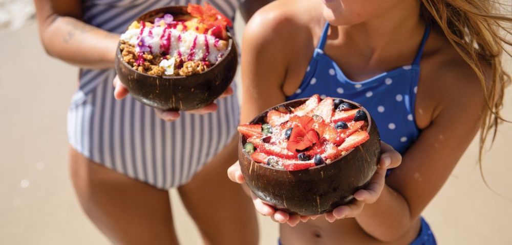 Two girls in swimsuits hold coconut bowls filled with colorful fruit and granola, showcasing a sunny beach day.