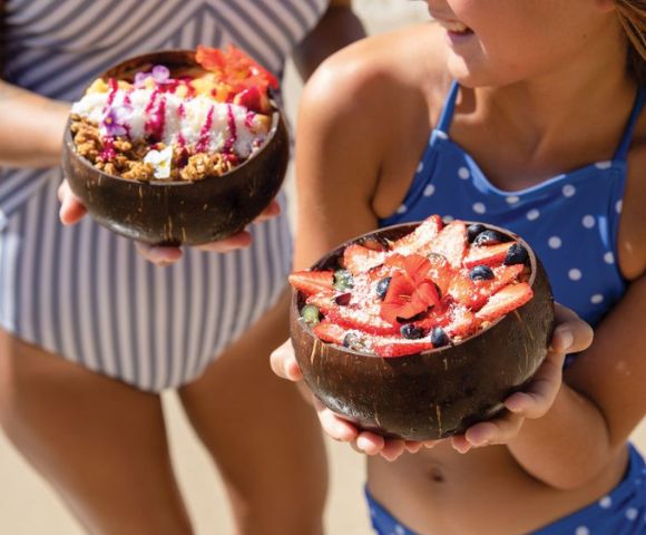 Two girls in swimsuits hold coconut bowls filled with colorful fruit and granola, showcasing a sunny beach day.