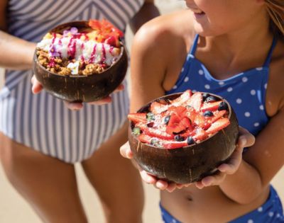 Two girls in swimsuits hold coconut bowls filled with colorful fruit and granola, showcasing a sunny beach day.