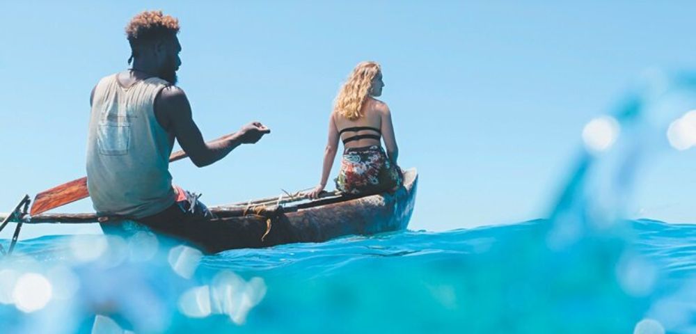 A woman and a man sit in a canoe on bright blue ocean water, with clear skies above, surrounded by gentle waves.