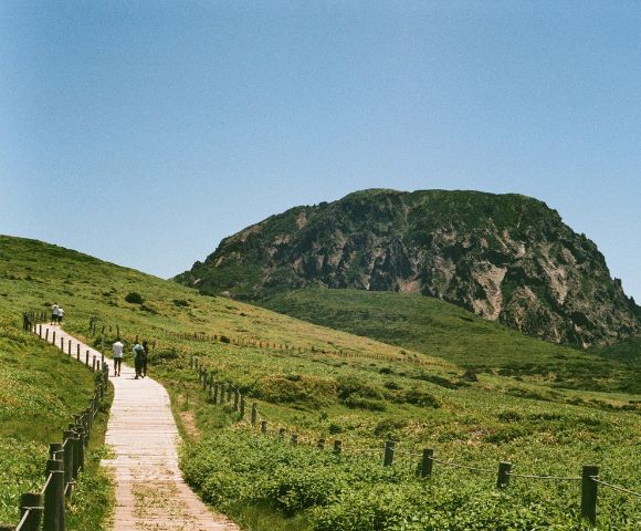 A serene landscape featuring a wooden walkway winding through lush green fields towards a rocky hill under a clear blue sky. People stroll along, enjoying nature.