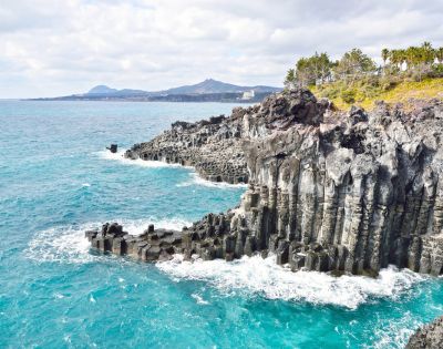 Stone statues of divers stand on a sandy beach with scattered rocks. The bright blue sea and a clear sky create a serene, tranquil atmosphere.