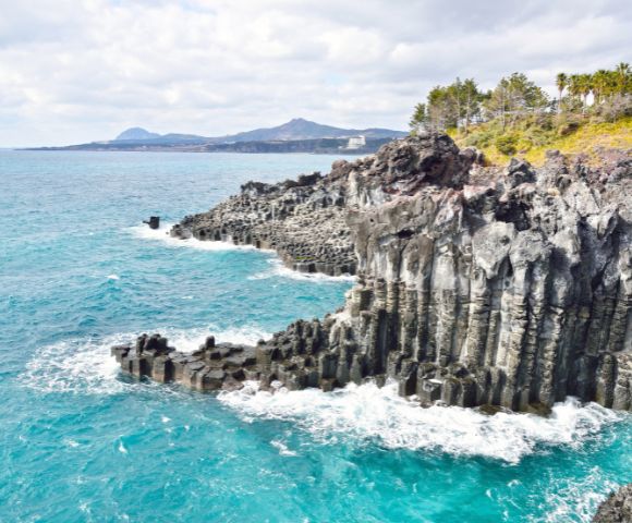 Stone statues of divers stand on a sandy beach with scattered rocks. The bright blue sea and a clear sky create a serene, tranquil atmosphere.