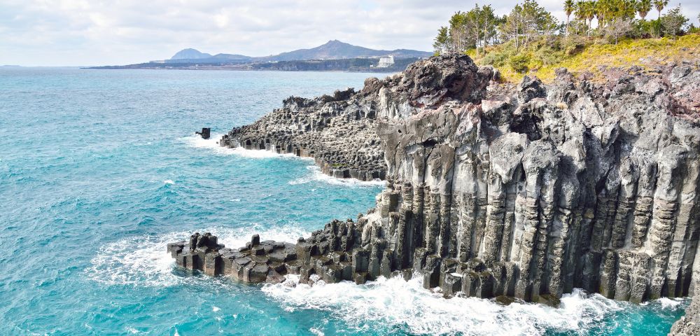 Stone statues of divers stand on a sandy beach with scattered rocks. The bright blue sea and a clear sky create a serene, tranquil atmosphere.